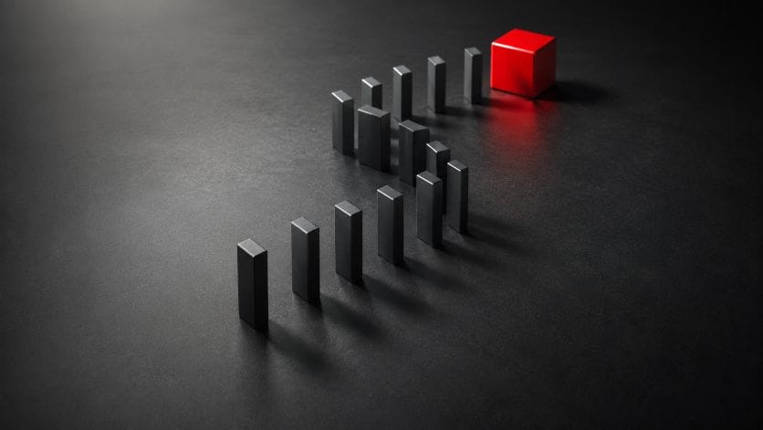 A line of black rectangular dominoes forming an S-shape curve stands on a dark, textured surface, leading towards a single red cube at the far end.